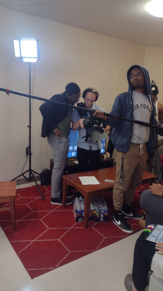 In the foreground is a man holding a boom mic. Behind him is a man looking over another man's shoulder watching the screen of a large film camera