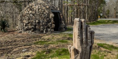 Carved praying hands holding a barbed wire cross, in front of a rock hut.