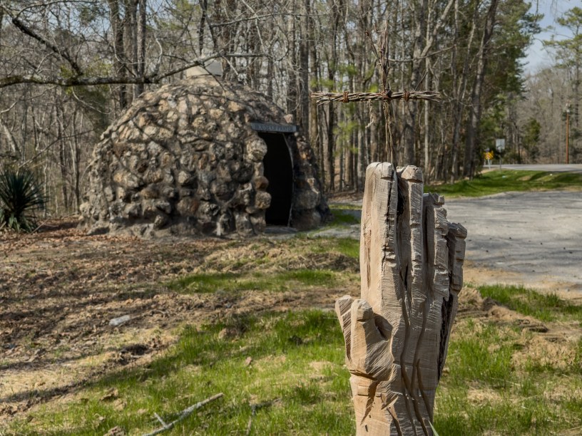 Carved praying hands holding a barbed wire cross, in front of a rock hut.
